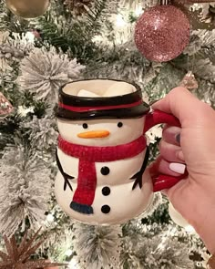 a hand holding a snowman mug in front of a christmas tree