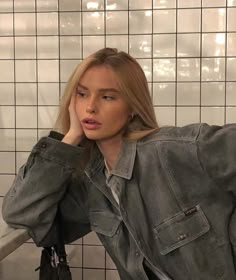 a woman sitting in front of a tiled wall with her hand on her head and looking at the camera