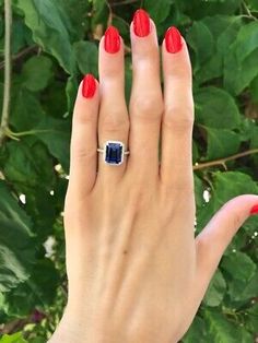 a woman's hand with red nail polish holding an engagement ring in front of green leaves