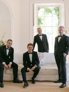 Seated black tie groom leaning forward on his knees, surrounded by his groomsmen, all bathing in natural window light. Elegant bride in a Suzanne Neville wedding dress with high lace neck, long lace sleeves and large bow at the back. Katie Julia Photography. Black Tie Groom, Suzanne Neville Wedding Dress, Suzanne Neville Wedding Dresses, On His Knees, Long Lace Sleeves, Suzanne Neville