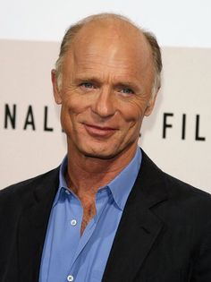 an older man in a blue shirt and black blazer smiles at the camera while standing next to a white wall