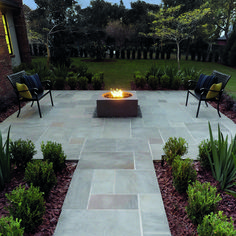 a fire pit in the middle of a patio surrounded by plants and trees, with chairs around it