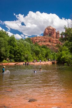 people are wading in the water near some red rocks and green trees on a sunny day