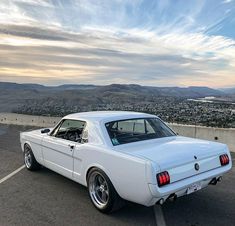 a white mustang parked in a parking lot with mountains in the back ground and clouds in the sky