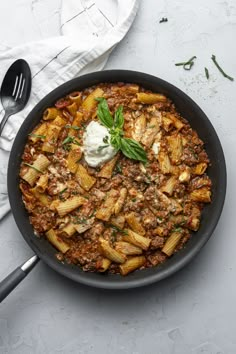 a skillet filled with pasta and meat on top of a white table next to utensils