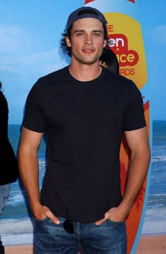 a man standing in front of a surfboard on the beach with his hands in his pockets