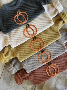 five sweaters with pumpkin designs on them sitting on a wooden floor, all in different colors