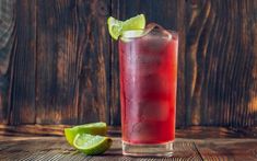 a tall glass filled with red liquid and lime wedges next to it on a wooden table