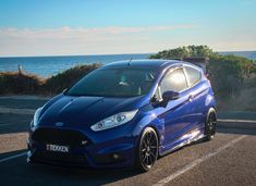 a blue car is parked on the side of the road near the ocean and trees