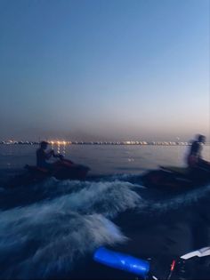 two people riding jet skis on top of a body of water at night with city lights in the background