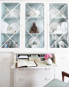 a white china cabinet with glass doors and plates on it's front, sitting next to a chair