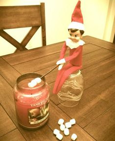 an elf sitting on top of a wooden table next to a jar filled with marshmallows