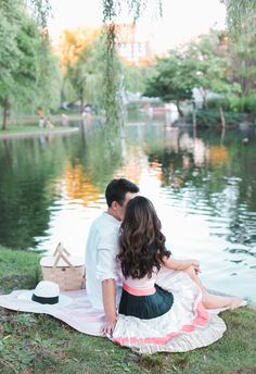 a man and woman sitting on top of a blanket next to a lake in the park