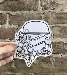 a hand holding up a sticker with a helmet and flowers on it in front of a brick wall