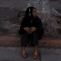 a woman with headphones sits on the curb next to a tree and looks off into the distance