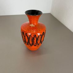 an orange and black vase sitting on top of a gray table next to a white wall