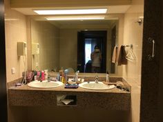 a bathroom with two sinks and a mirror in it's corner, next to a woman taking a selfie