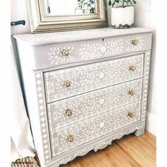 a white dresser with gold handles and drawers in front of a mirror on top of a wooden floor