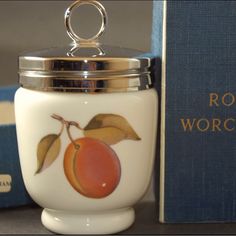 a glass jar with a fruit painted on it next to a blue book and silver lid