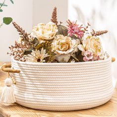 a white rope basket with flowers in it on a wooden table next to a potted plant