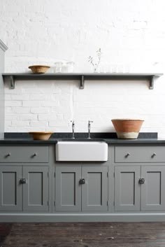 a kitchen with gray cabinets and white brick wall, two sinks and open shelving