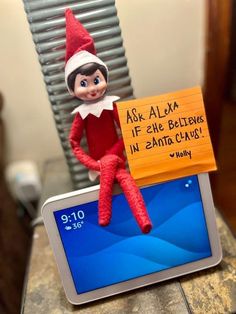 an elf sitting on top of a tablet computer next to a sign that says ask alex if she believe in santa claus