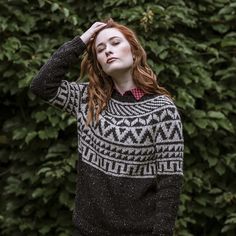 a woman with red hair wearing a black and white sweater standing in front of some bushes