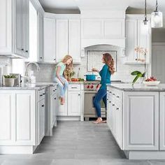 two women in a kitchen with white cabinets and gray flooring, one standing at the sink