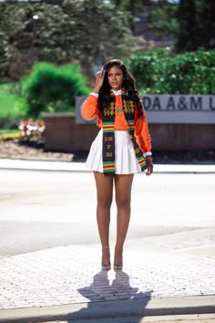 a woman in an orange jacket and white dress is standing on the sidewalk with her hands behind her head