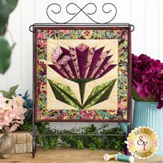 a quilted wall hanging with purple flowers and greenery in front of the frame