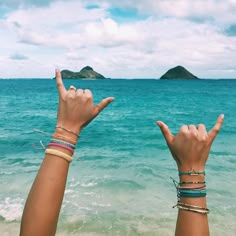 two hands holding up the peace sign in front of an ocean with mountains and blue sky