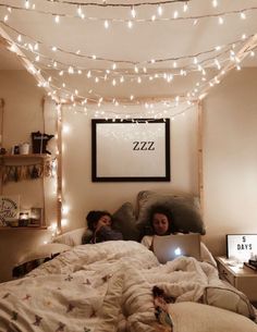 two people are laying in bed with lights strung above them and one person is using a laptop