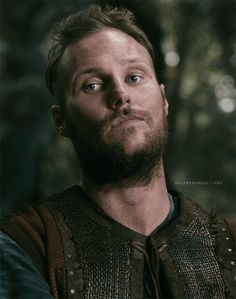 a man with a beard wearing a brown vest and looking into the distance while standing in front of trees