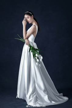 a woman in a white wedding dress holding flowers and looking at her cell phone while standing against a dark background