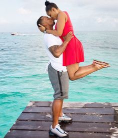 a man and woman kissing on a dock in front of the ocean with blue water