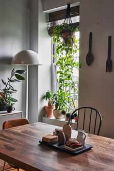 a wooden table topped with plates and cups next to a window filled with potted plants