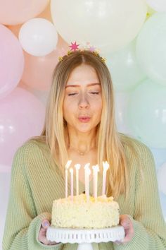 a woman holding a cake with candles in front of her face and balloons behind her