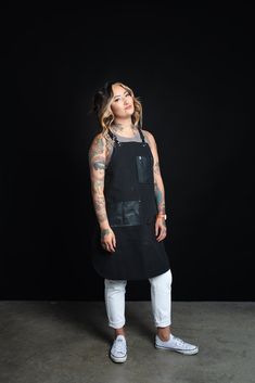 a woman standing in front of a black background wearing an apron and white pants with tattoos on her arms