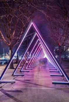 a group of people walking down a sidewalk next to trees and benches with neon lights on them