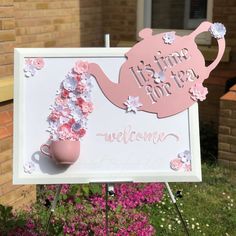 a welcome sign with pink flowers and a teapot on it in front of a brick building