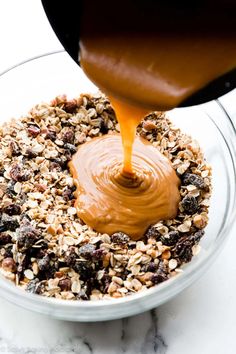 someone pouring caramel sauce on top of granola in a glass bowl with white marble countertop