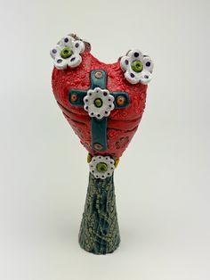 a red heart shaped vase with flowers on it's side and a cross in the middle