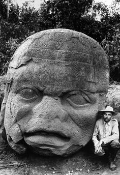 a man sitting next to a large stone statue with a face on it's head