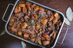 meat and potatoes in a roasting pan on top of a wooden table with leaves