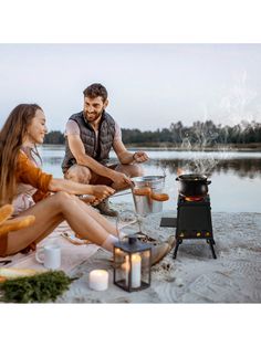 a man and woman sitting on the beach next to a campfire with hot dogs