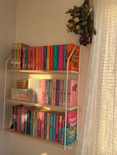 a book shelf filled with lots of books next to a white curtain and flowers on the window sill