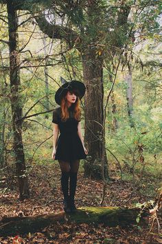 a woman in a black dress and hat stands on a log in the woods with trees behind her