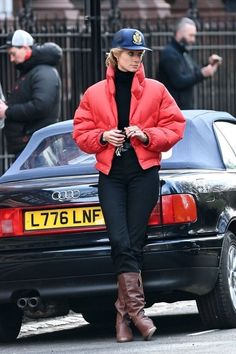 a woman in a red jacket and black pants is walking past a car on the street