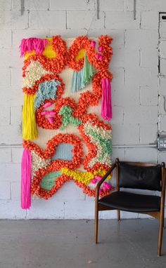 a chair sitting in front of a wall hanging with tassels and flowers on it