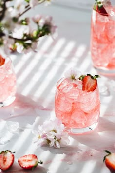 two glasses filled with ice and strawberries on top of a white table next to flowers